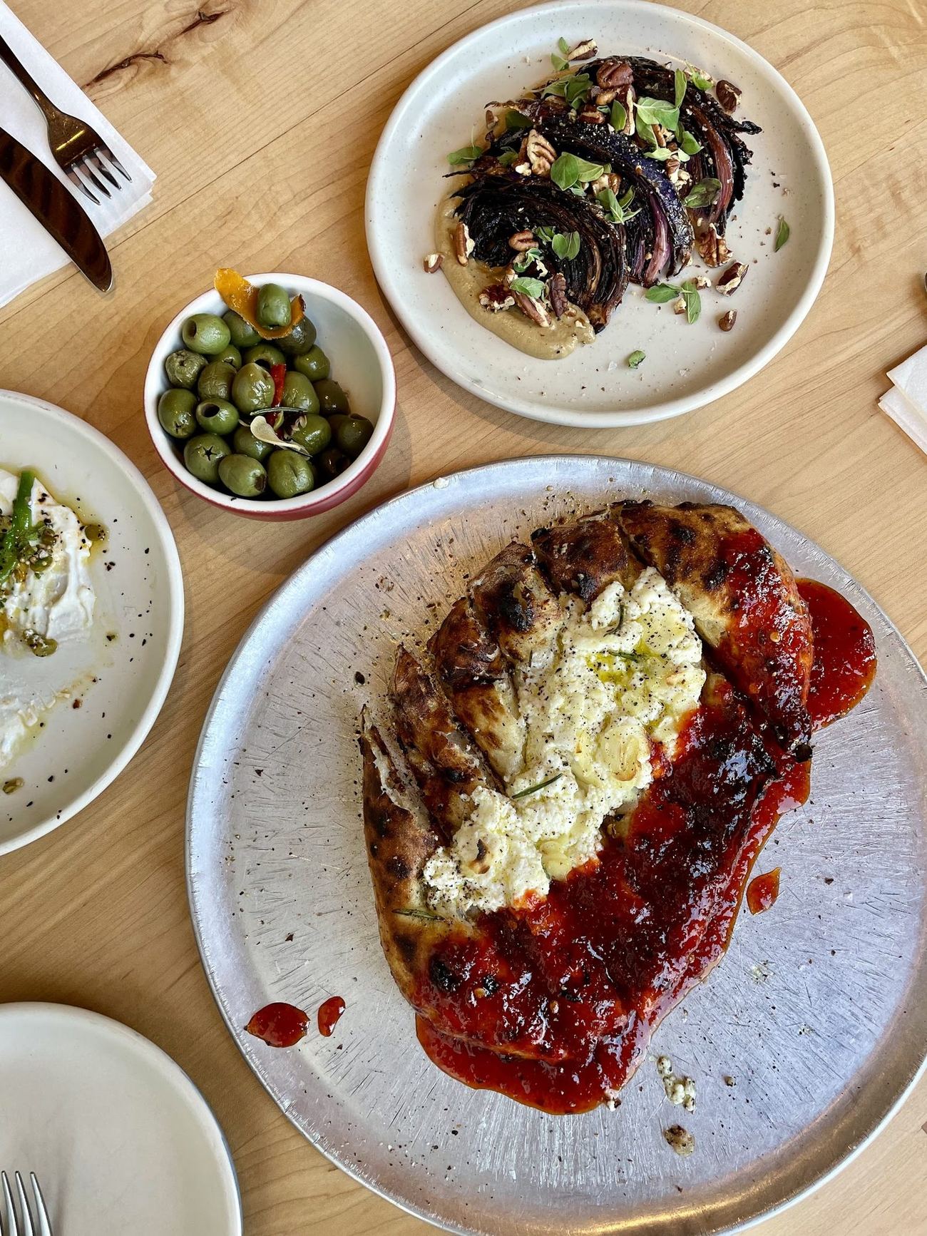 A table of small plates: a beetroot and herb salad, a bowl of olives and a slice with red sauce, on pale ceramic plates