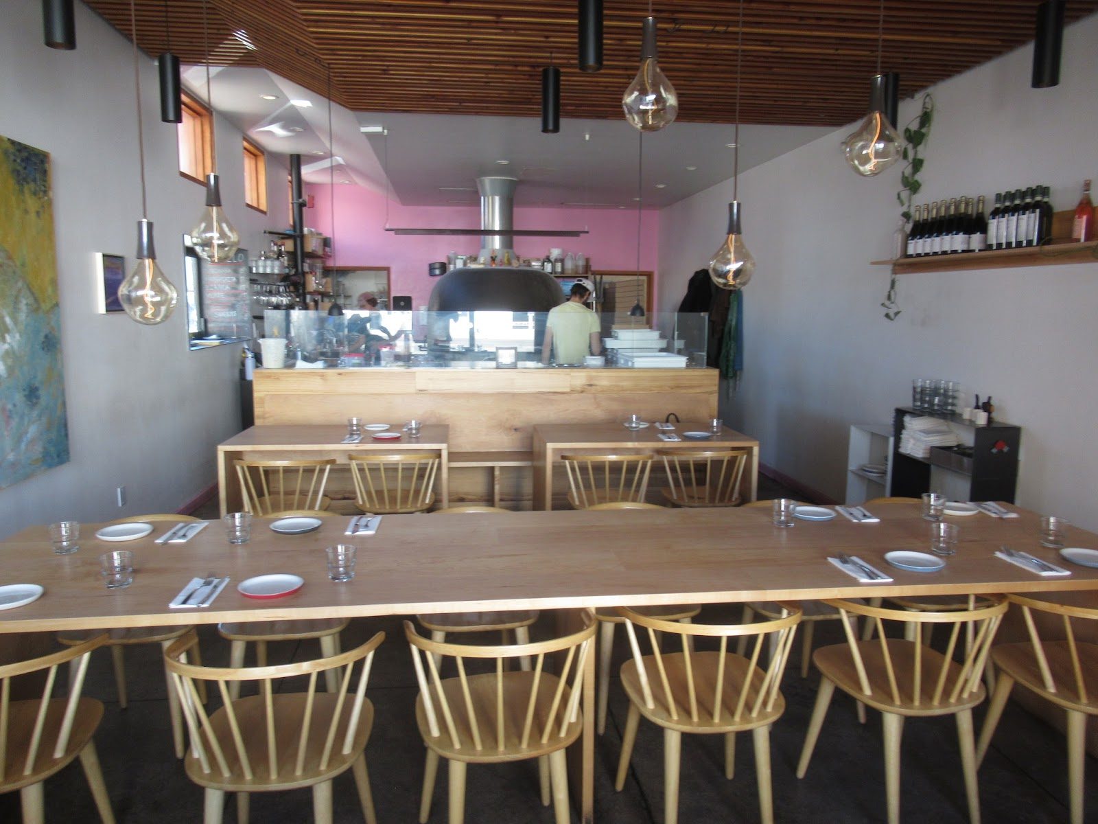 The dining room: a long communal wood table with pale spindle chairs, pendant lights, and an open kitchen beyond a pink wall